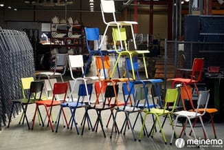 Kleurrijke stapelstoelen in rood, blauw, geel, oranje en wit met zwarte metalen frames in een magazijnruimte.