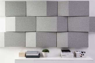 Grijze akoestische wandpanelen in verschillende tinten op witte wand boven witte plank met boeken, plant en houten kubus.