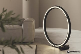Ovaal verlichte spiegel met zwart frame en LED-verlichting, staand op zwarte voet, naast beige poef en plant op houten tafel.