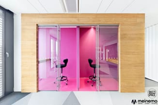 Twee glazen kantoorboxen met roze wanden, houten frame, zwarte bureaulstoelen, in moderne werkomgeving.