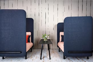 Twee donkerblauwe akoestische belcellen met roze zitgedeelten, zwarte bijzettafel met bloemen, houten vloer en witte houten w