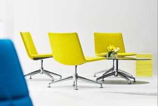 Drie gele kantoorstoelen met zilveren onderstel rond een kleine tafel met bloemen en boeken, naast een blauwe stoel op witte