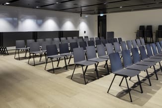 Moderne vergaderruimte met rijen grijze stoelen met zwart metalen frame op houten vloer, witte wanden en zwart plafond.