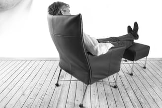 Man in wit overhemd zit ontspannen in zwarte leren fauteuil met voetenbankje op houten vloer.