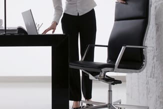 Kantoor met zwarte bureau, zilveren bureaustoel en laptop bij raam; persoon in wit overhemd en zwarte broek aan het werk.