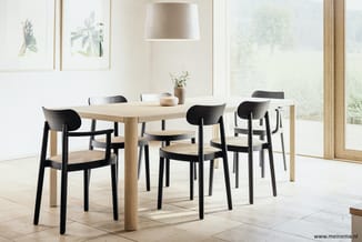 Lichte eetkamer met lichte houten tafel, zwarte stoelen met riet, hanglamp, wanddecoratie en grote ramen.