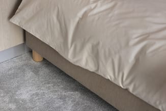 Grijs bed met beige beddengoed en houten poot, tegen grijze vloer en beige wand.