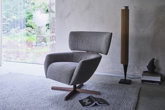 Grijze draaisstoel met hoge rugkussen en houten kruisvoet, geplaatst op grijs tapijt in minimalistische betonnen ruimte met r