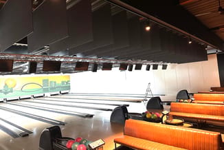 Bowlingbaan met oranje gestoffeerde zitbanken, zwarte bowlingballen en witte lanes onder industrieel plafond met zwarte akoes