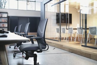 Modern kantoor met zwarte bureaustoel op wielen, witte vergadertafel, grijze stoelen en glazen scheidingswand, boekenkasten,
