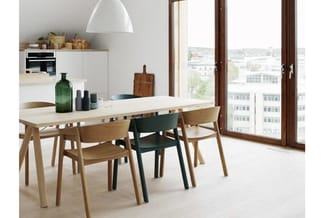 Moderne eetkamer met houten vergadertafel, houten en groene stoelen, witte hanglamp, keukenblok op achtergrond en grote ramen