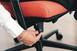 Hand bedient armsteun van rode bureaustoel met zwart frame en wielenonderstel.