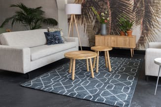 Moderne woonkamer met grijze linnen bank, twee houten bijzettafels, blauwe geometrische vloerkleed, houten dressoir, staanlam