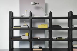 Zwarte modulaire kastenwand met vakken gevuld met boeken, gele dozen en kantoorartikelen. Witte bureaulamp bovenop. Minimalis