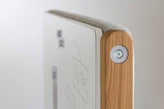 Hoekdetail van wit paneel met houten rand en ronde metalen ventilatieopening, tegen grijze achtergrond.