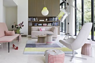 Modern woonkamer met beige sofa, grijze fauteuil, roze zitbank, houten kasten met boeken, geel hangpendel, kleurig vloerkleed