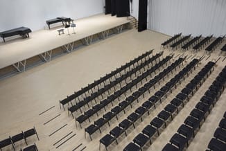 Zaalsetting met zwarte stoelen in rijen opgesteld, verhoogd podium met vleugels en microfoonstandaard, witte wanden en spotve
