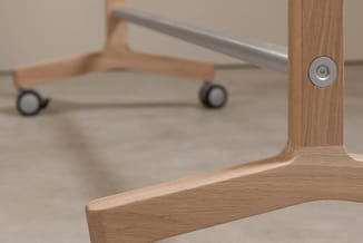 Houten garderobe met bovenliggende rail en twee zwarte wielen, voorzien van een metalen bevestigingspunt op de zijkant.