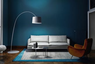 Moderne woonkamer met grijze hoekbank, cognackleurige fauteuil, boogvloerlamp en blauwe muurschildering.