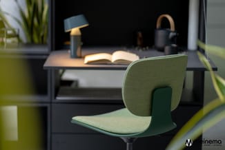 Groene stoel met gestoffeerde zitting en rugleuning aan bureau met open boek, lamp en plantjes in moderne kantooromgeving.