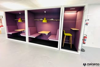 Drie akoestische belcellen met paarse binnenbekleding, geel hangend armatuur, houten werkbladen en gele barkruk in kantoorrij