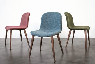 Drie gestoffeerde stoelen in roze, blauw en olijfgroen met houten poten, op betonvloer tegen witte wand.