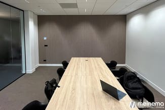Moderne vergaderruimte met lichte houten vergadertafel, zwarte stoelen, verticale bruine wandbekleding en glazen wand.