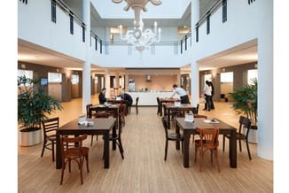 Kantoorruimte met houten eettafels, houten stoelen, groene planten, bruine vloer, witte plafonds met kroonluchter en personee