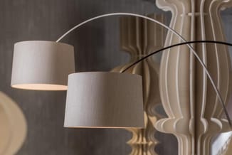 Boogvloerlamp met twee witte stoffen kappen boven grijze muur met decoratief wit patroon.