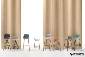 Acht barstoelen in verschillende kleuren en vormen tegen geribde houten wand. Zitoppervlakken in zwart, wit, blauw, oranje en