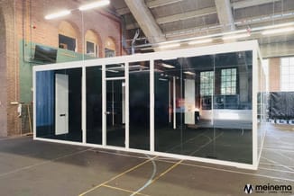 Moderne glazen belcel met wit aluminium frame en zwarte glaspartijen, geplaatst in industriële hal met bakstenen muur.