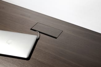 Grijsbruine houten tafel met ingebouwde zwarte rechthoekige stopcontact, wit apparaat links.