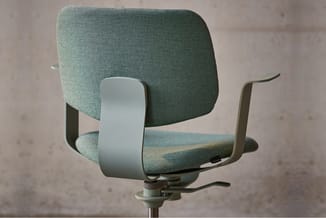 Grijsgroene bureaustoel met textiel bekleding, gebogen rugsteun, verstelbare armen en zwenkvoet, tegen betonnen achtergrond.