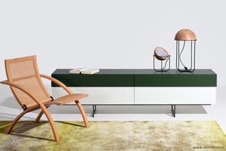 Houten loungestoel met rattan bekleding naast groen-wit sideboard met houten lamps en open boek op geel-groen tapijt.