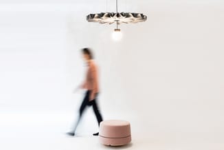Modern hanglamp met geribbelde metallic kap boven roze poef in kantoorruimte met bewegende persoon.