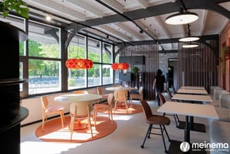 Modern kantoor met ronde tafel, oranje stoelen, rode hanglamp, grote ramen, zwarte balken, wit plafond en beige vloer.