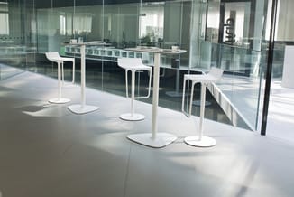 Moderne kantoorruimte met drie witte holle staafels en bijbehorende witte barkrukken langs glazen wand, met uitzicht op werkr