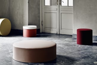 Cylindrische poefs en zitobjecten in beige, rood en grijs in een minimalistische ruimte met betonnen muren en dubbele deuren.
