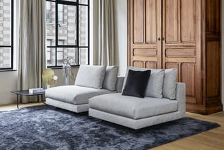 Grijze modulaire loungeset met chaise longue, kussens en zwart accent kussen in kantoor/woonruimte met houten kasten en raam.