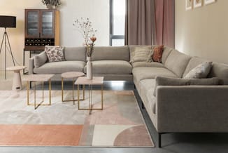 Grijze hoekbank met decoratieve kussens, salontafels met roze blad en gouden frame, geométrisch tapijt, staanlamp en wandkast