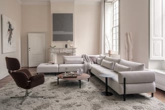 Moderne lounge met beige hoekbank, bruine fauteuil, rond tafelblad en grijze vloerkleed in lichte woonkamer met grote ramen.