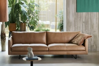 Bruine leren bank met vier zitplaatsen, metalen poten, in moderne woonruimte met grote ramen en groene planten.