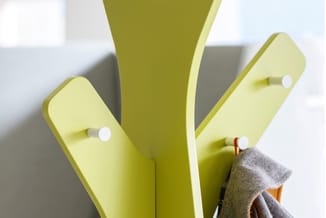 Gele kapstok met drie armen, uitgevoerd in modern design met witte knoppendetails. Grijs kledingstuk eraan opgehangen.