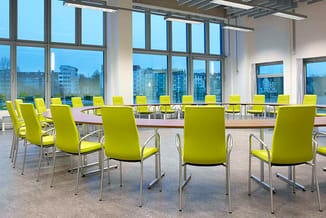 Moderne vergaderruimte met gele stapelstoelen met aluminium frames aan houten vergadertafels. Grote raampartijen met stadsuit