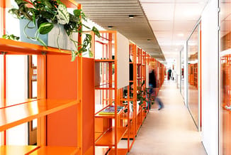 Moderne kantoorcorridor met rij oranje belcellen aan linkerkant, glaswanden, hangende planten, betonnen plafond en personen i