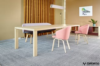 Vergaderruimte met lichte houten vergadertafel, roze stoelen met houten poten, witte en grijze stoelen, grijs tapijt, gele go