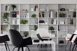 Moderne kantoorruimte met witte boekenkast, zwarte en wit-rode bureaustoelen, witte salontafel, planten en boeken.