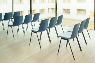 Blauwe kunststof stoelen met zwart metalen frame in rijen op lichte houten vloer, grote ramen en witte gevel op achtergrond.