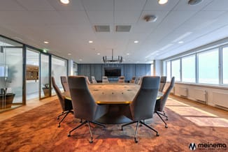 Moderne vergaderruimte met ovale houten tafel, grijze kantoorstoel en ronde basis, grote ramen en blauwe wandbekleding.