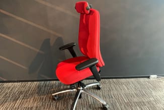 Rode ergonomische bureaustoel met hoofdsteun en zwarte armleuningen, op wielen, in kantoorruimte met grijze wand.